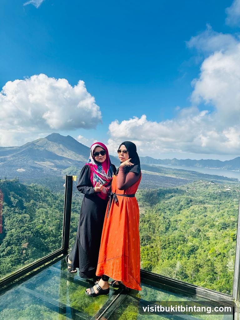 Two women posing on a glass viewing platform with a scenic mountain and lake backdrop.