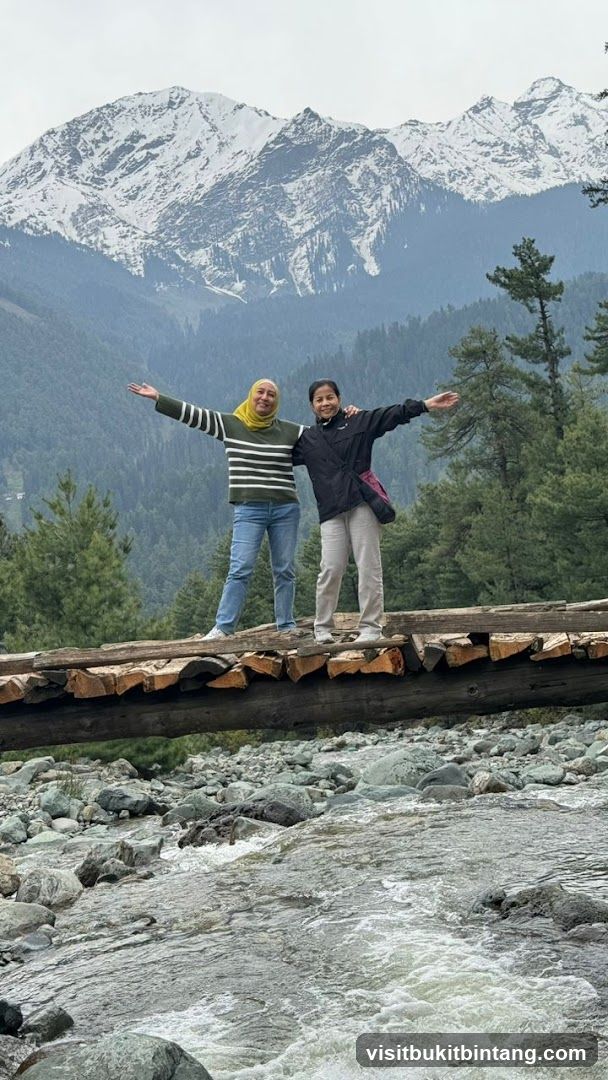 Two women on a wooden bridge over a stream with snowy mountains in the background