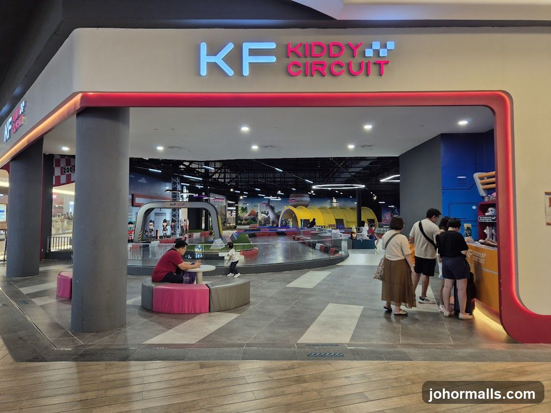 The entrance to an indoor amusement park for children. The sign above the entrance reads "KF KIDDY CIRCUIT" in blue and pink neon letters. Inside, there is a go-kart track with colorful barriers and cartoon-like decorations. Several children and adults are visible inside, some near the track and others at a counter.