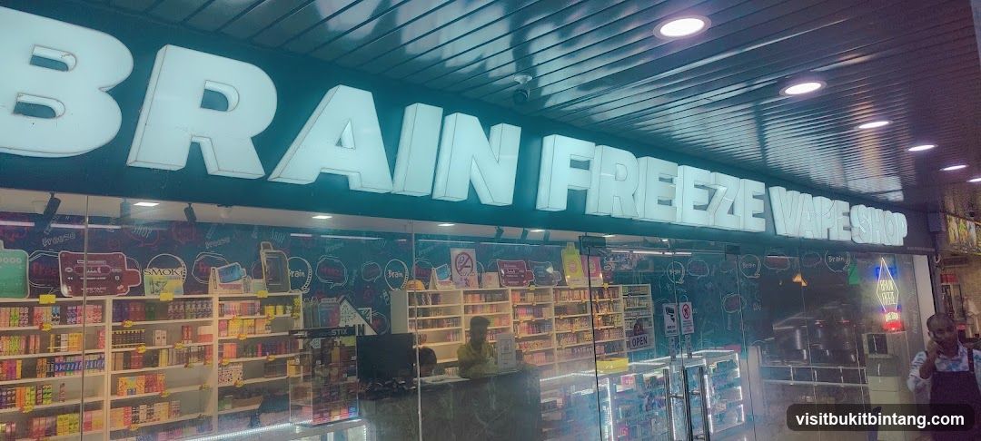 The exterior of a vape shop with large illuminated signage that reads "BRAIN FREEZE VAPE SHOP". The storefront features glass doors and windows displaying shelves stocked with various vape products. A person is visible behind a counter inside the shop.