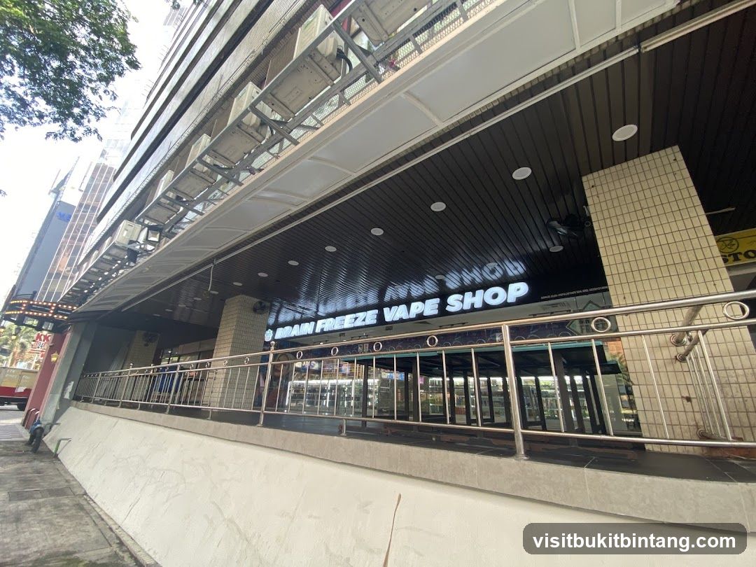 The exterior of a vape shop with a prominent illuminated sign that reads "BRAIN FREEZE VAPE SHOP". The shop has large glass windows and doors, reflecting the interior. A metal railing runs along the front of the shop.