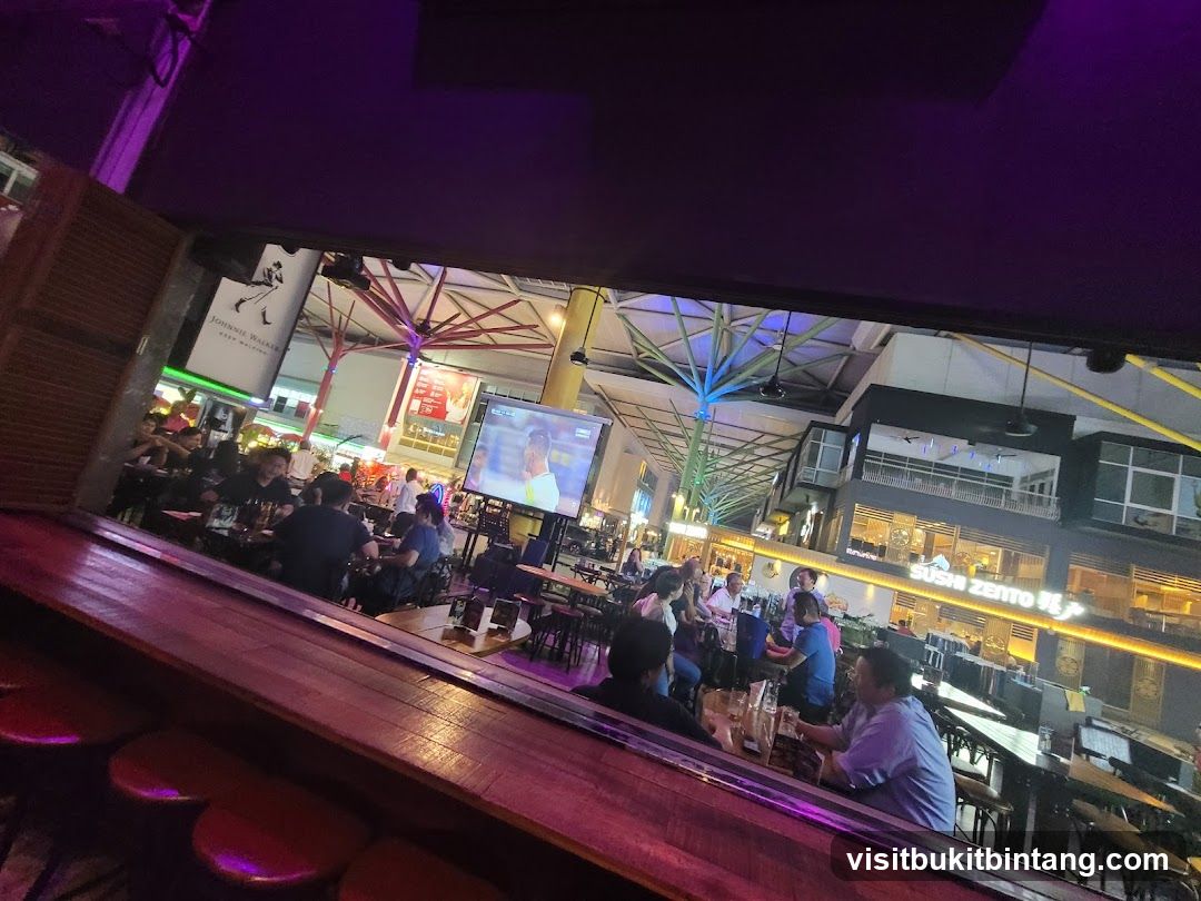 People dining at tables in a lively outdoor area with screens showing sports