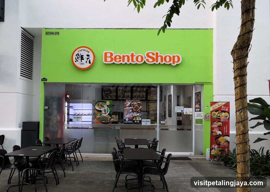 A bright green storefront with 'Bento Shop' signage and a circular logo featuring Japanese characters. The entrance has glass doors and windows displaying menu boards inside. Outdoor seating is arranged on the left side, while promotional posters stand near the right entrance.