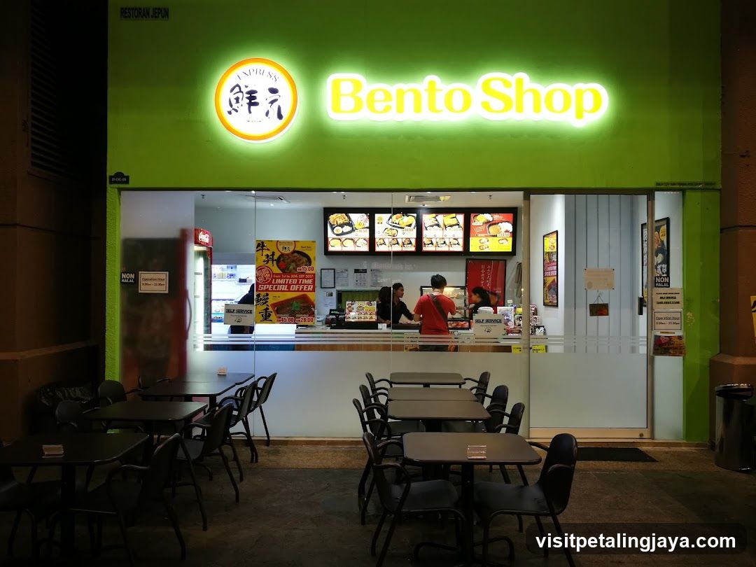 The image shows the exterior of a brightly lit bento shop with green walls and illuminated signage reading 'Bento Shop' in yellow letters alongside a circular logo featuring Japanese characters. Outside seating is arranged on the sidewalk, while inside, staff are visible behind the counter serving customers. Menu boards display various food items above the service area.