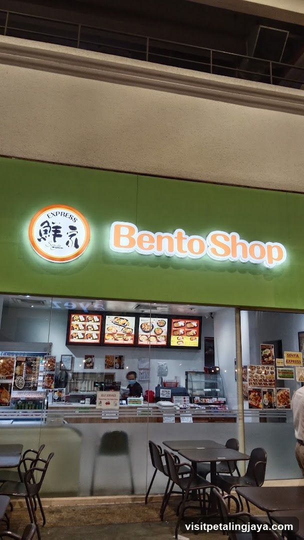 A storefront of a Bento Shop with green signage and illuminated 'Bento Shop' text alongside an orange circular logo featuring Japanese characters. The shop has glass windows displaying menu boards inside, showing various bento box options. Outside seating is visible with tables and chairs in front.