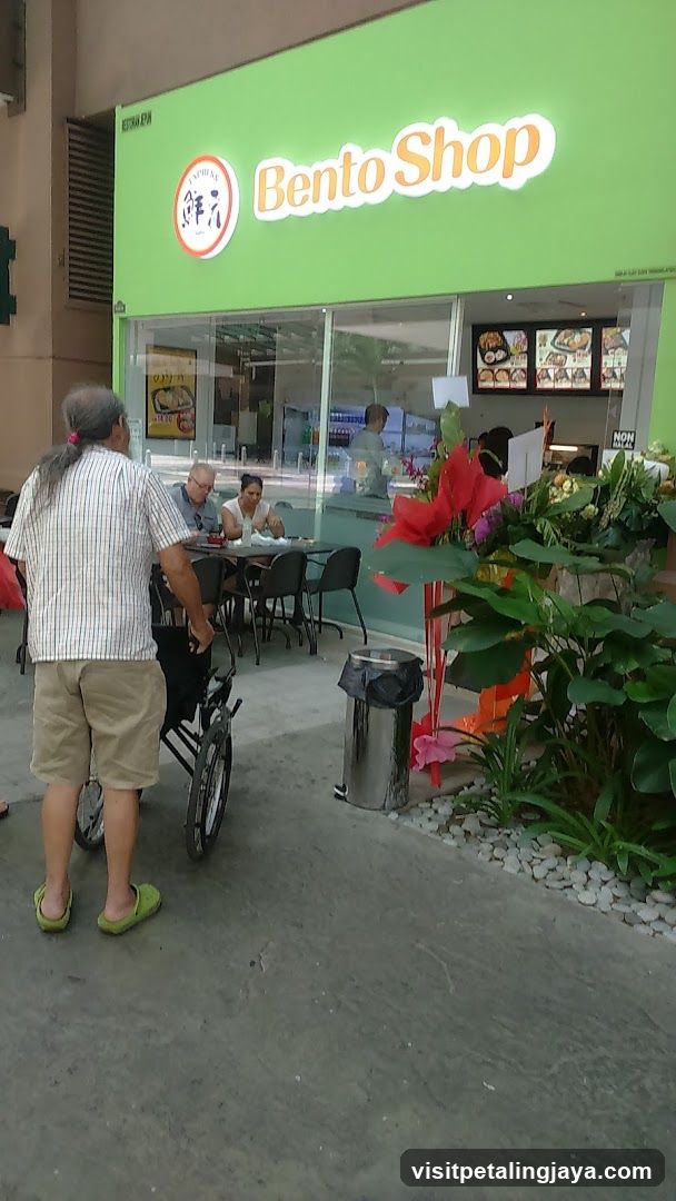 A green storefront with 'Bento Shop' signage and a circular logo featuring Japanese characters. A person in a wheelchair is outside, while others sit at tables near the entrance. Decorative plants and flowers are placed along the sidewalk.