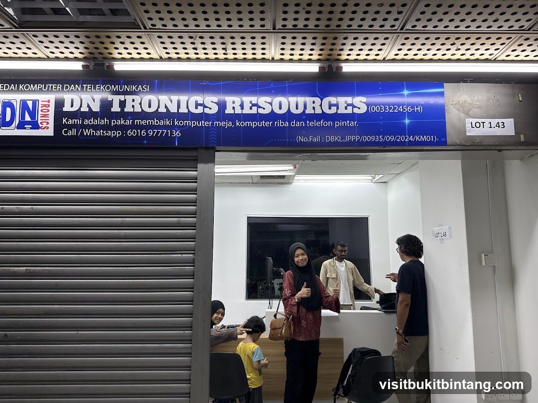 A shop front with a large blue sign above the entrance that reads "DN TRONICS RESOURCES". Below the name, it states "Kami adalah pakar membaiki komputer meja, komputer riba dan telefon pintar." and provides a call/whatsapp number. The entrance has a grey roller shutter partially open, revealing people inside.