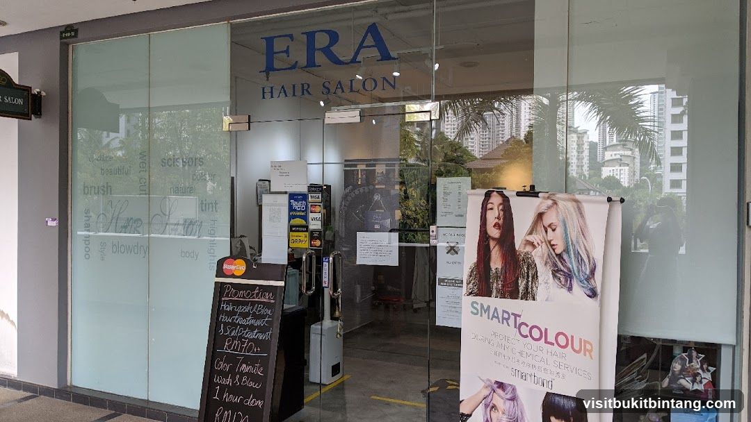 The exterior of ERA Hair Salon features glass doors with frosted panels displaying various hair-related terms. The salon's name, "ERA HAIR SALON," is prominently displayed above the entrance. A promotional blackboard lists services and prices, including a "Promotion Hair-upstu Blow Hurtreatment & Scalptatnunt" for RM70. Another offer is for "Color Tminute Wash & Blow 1 hour done" for RMDA. A large banner showcasing "SMART COLOUR" and hair models is placed to the right of the entrance. Payment options like Visa, Mastercard, and GR PAY are visible.