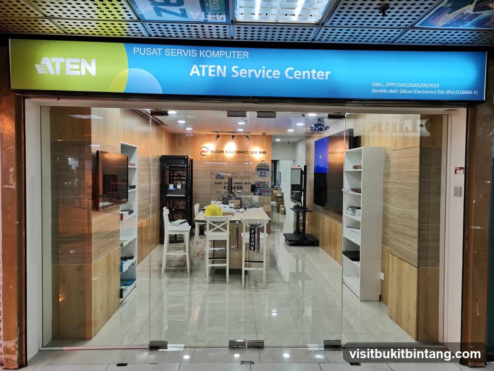 The storefront of ATEN Service Center, a computer service center. The entrance features a large glass facade with a prominent blue and green sign above that reads "ATEN PUSAT SERVIS KOMPUTER ATEN Service Center". Inside, the shop appears to be an electronics repair and service center with shelves, tables, and computer equipment visible.
