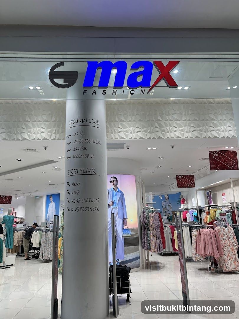 The image shows the entrance to a Max Fashion store. The store's logo, "max FASHION", is prominently displayed above the entrance. A large pillar in the foreground has handwritten signs indicating the departments on the ground and first floors, such as Ladies, Lingerie, Accessories, Mens, and Kids Footwear. The interior of the store is visible, with racks of clothing and mannequins displaying outfits. Sale signs are also visible within the store.