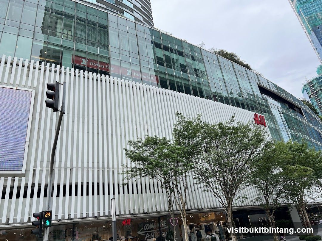 A modern building facade with a prominent H&M logo in red. The building features a white vertical slatted design on its lower levels and a glass-covered upper section. Trees line the sidewalk in front of the store. A traffic light is visible to the left.