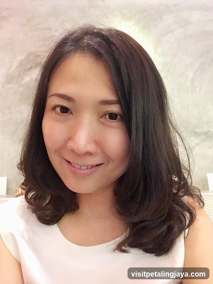 Portrait of a woman with shoulder-length dark hair smiling gently at the camera, wearing a white sleeveless top against a light textured wall background.