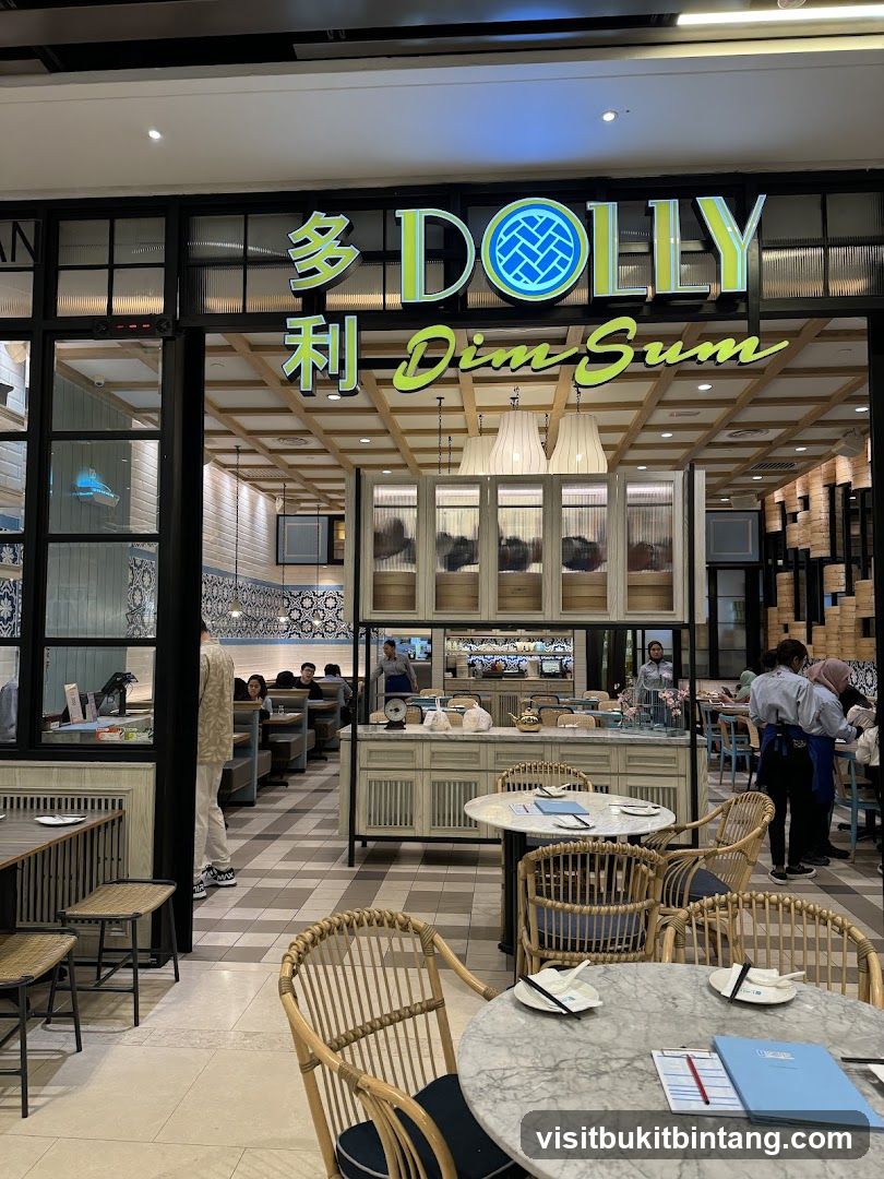 The exterior of a dim sum restaurant with a prominent sign that reads "DOLLY Dim Sum" in English and Chinese characters. The entrance features large windows and an open view into the dining area, which is decorated with light-colored furniture and traditional Chinese-inspired elements. Several tables are set with place settings, and some patrons are visible inside.