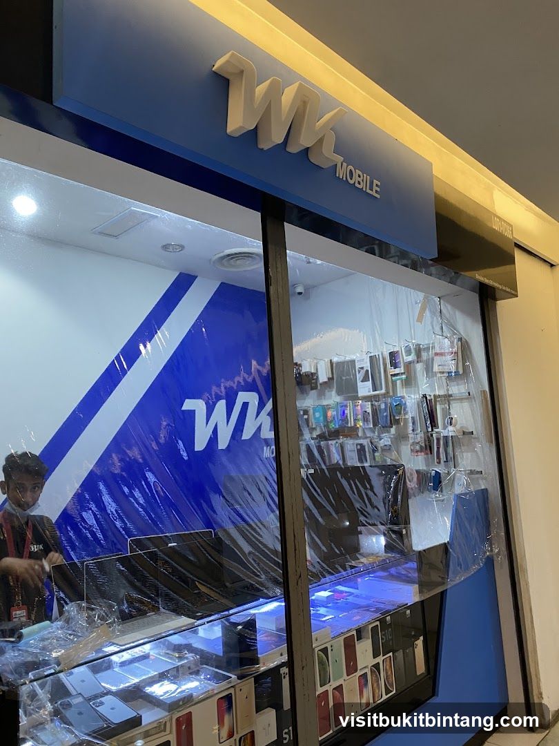 A storefront for a mobile phone shop named WK Mobile. The shop has a blue facade with the WK Mobile logo prominently displayed. Inside, various mobile phones and accessories are displayed in glass cases. A staff member wearing a mask is visible behind the counter.