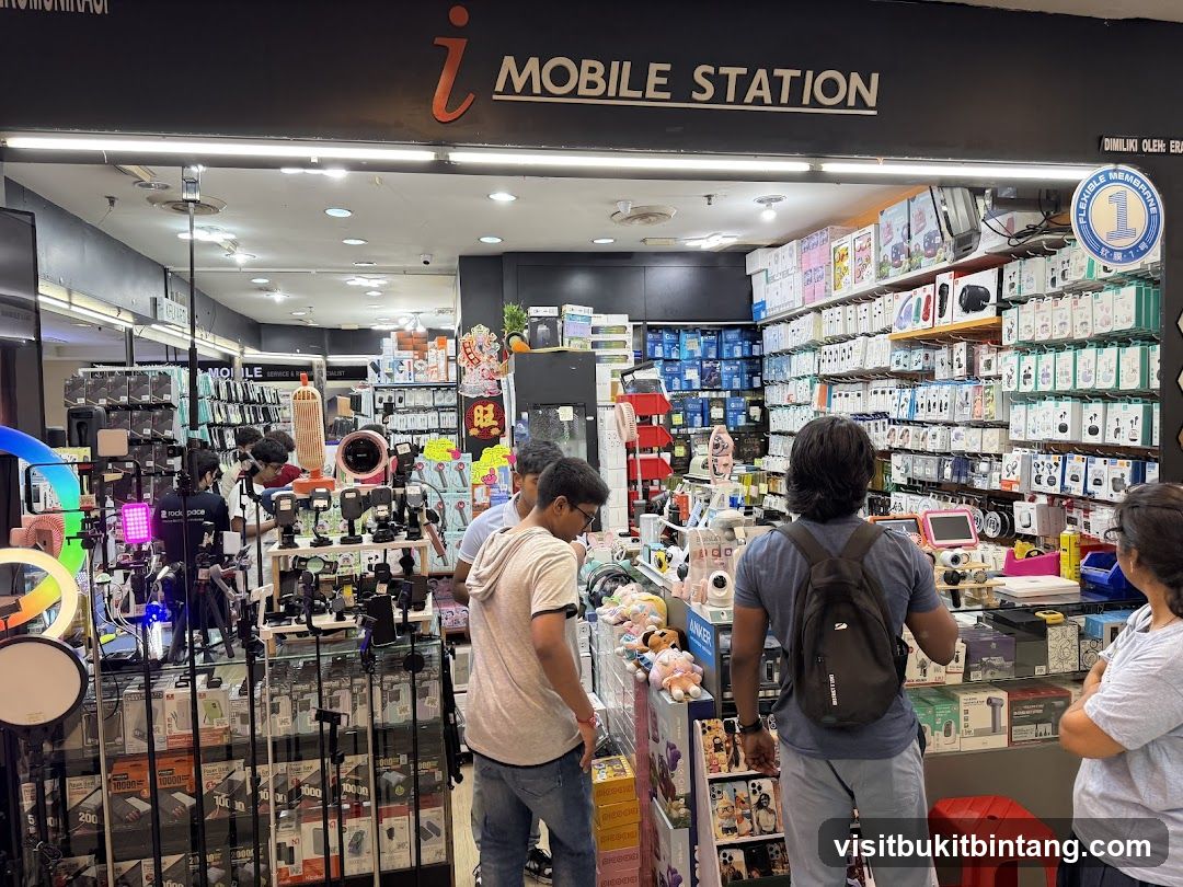 The exterior of a mobile accessories store called "i MOBILE STATION". The store is filled with various mobile accessories, including phone cases, chargers, headphones, and other electronic gadgets. Several customers are browsing the merchandise. The store's signage is clearly visible above the entrance.