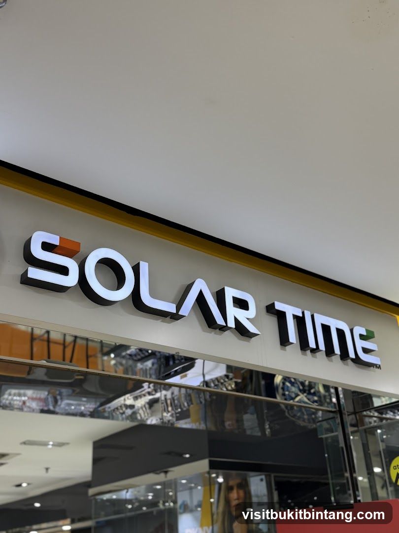 A close-up, angled shot of the illuminated sign for "SOLAR TIME" mounted above a store entrance. The sign features white, backlit letters with black trim. Below the sign, a reflective surface mirrors the interior of the mall, showing displays of watches and other merchandise. A portion of a poster featuring a woman's face is visible on the right.