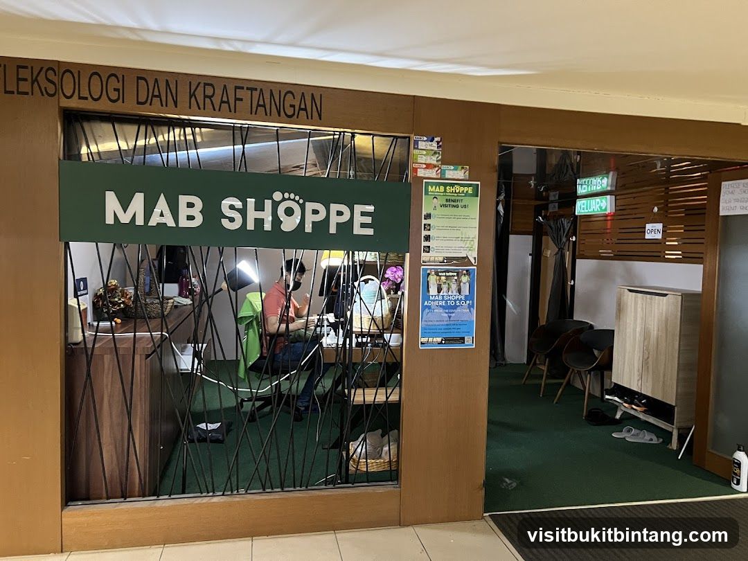 The storefront of MAB SHOPPE, a business offering reflexology and hand therapy services. The entrance features a green sign with white lettering that reads "MAB SHOPPE" and a foot graphic. Above the entrance, wooden lettering spells out "REFLEXOLOGY AND HANDICRAFTS". Inside, a person is seated at a desk with a computer, and there are various items on shelves. To the right, there is an open doorway with "OPEN" signage and a shoe rack.