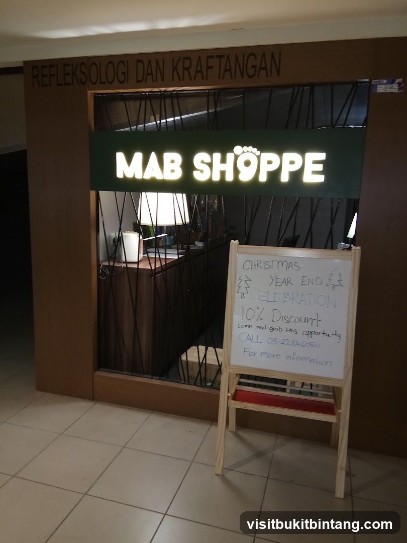 The storefront of MAB SHOPPE, a reflexology and handicraft business. The shop has a prominent illuminated sign with the business name. Above the sign, text reads "REFLEKSOLOGI DAN KRAFTANGAN". A whiteboard easel is placed in front of the shop, advertising a Christmas Year End Celebration with a 10% discount and a contact number.