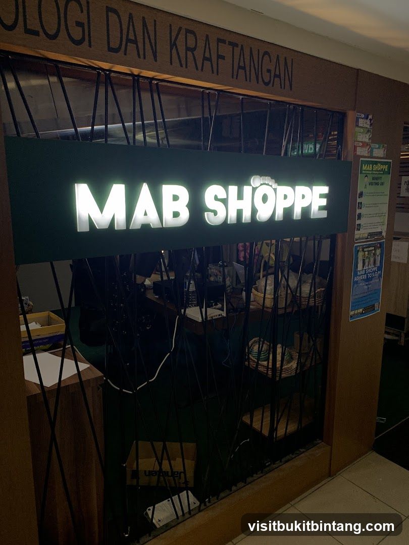 The exterior of a shop with a prominent illuminated sign that reads "MAB SHOPPE". Above the sign, wooden lettering spells out "LOGI DAN KRAFTANGAN". The shop entrance is partially obscured by a metal grate. To the right of the entrance, there are several posters and notices displayed on the wall.