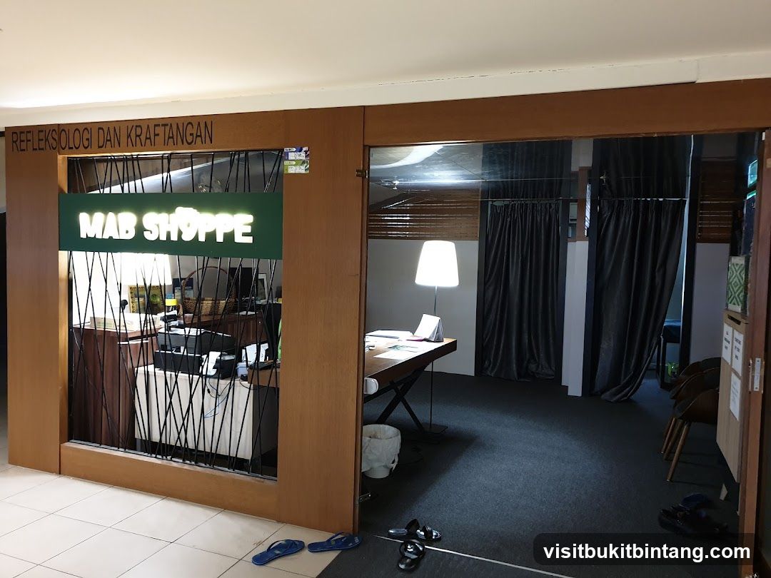 The entrance to Mab Shoppe is visible, with a green illuminated sign displaying the name. Above the sign, text reads "REFLEKSOLOGI DAN KRAFTANGAN". The interior of the shop is partially visible, showing a reception desk with a computer and a basket. To the right, a room with dark curtains and a lamp is seen.
