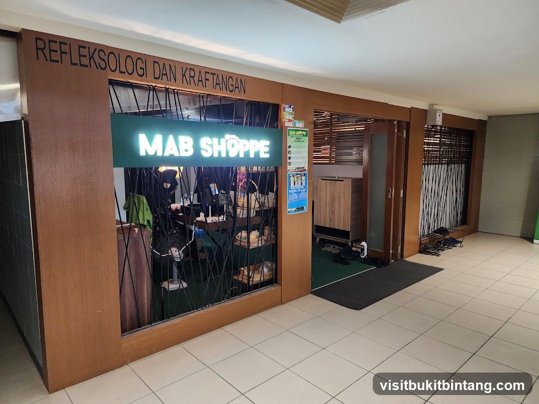 The exterior of a shop called Mab Shoppe, which offers reflexology and handcraft services. The shop has a wooden facade with a large glass window displaying various items. Above the window, the words "REFLEKSOLOGI DAN KRAFTANGAN" are visible. Inside, there are shelves with products, a fan, and a counter. A green sign with the shop's name, "MAB SHOPPE," is illuminated. To the right, there is an entrance with a wooden door and a sign indicating it is open.