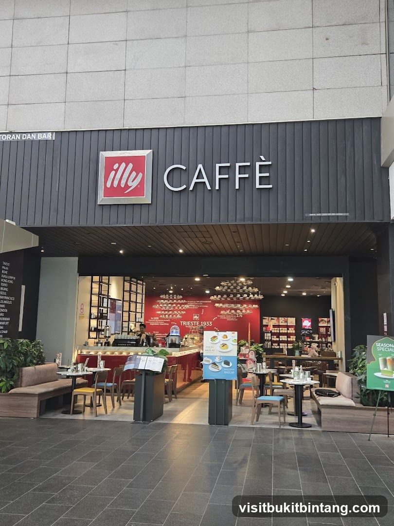 The exterior of an Illy Caffè coffee shop. The prominent signage above the entrance reads "illy CAFFÈ". The entrance opens to reveal a brightly lit interior with a red counter, seating areas, and displays. Tables and chairs are set up both inside and outside the shop. A seasonal special poster is visible to the right.