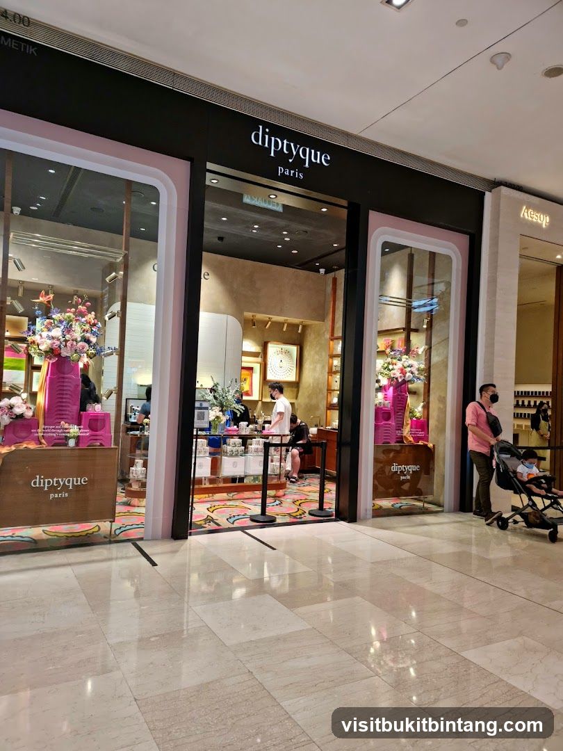 The exterior of a Diptyque Paris store, featuring large display windows showcasing floral arrangements and products. The store's name, 'diptyque paris', is prominently displayed above the entrance. The entrance is open, with a glimpse of the interior and staff. To the right, the adjacent store 'Aesop' is visible.