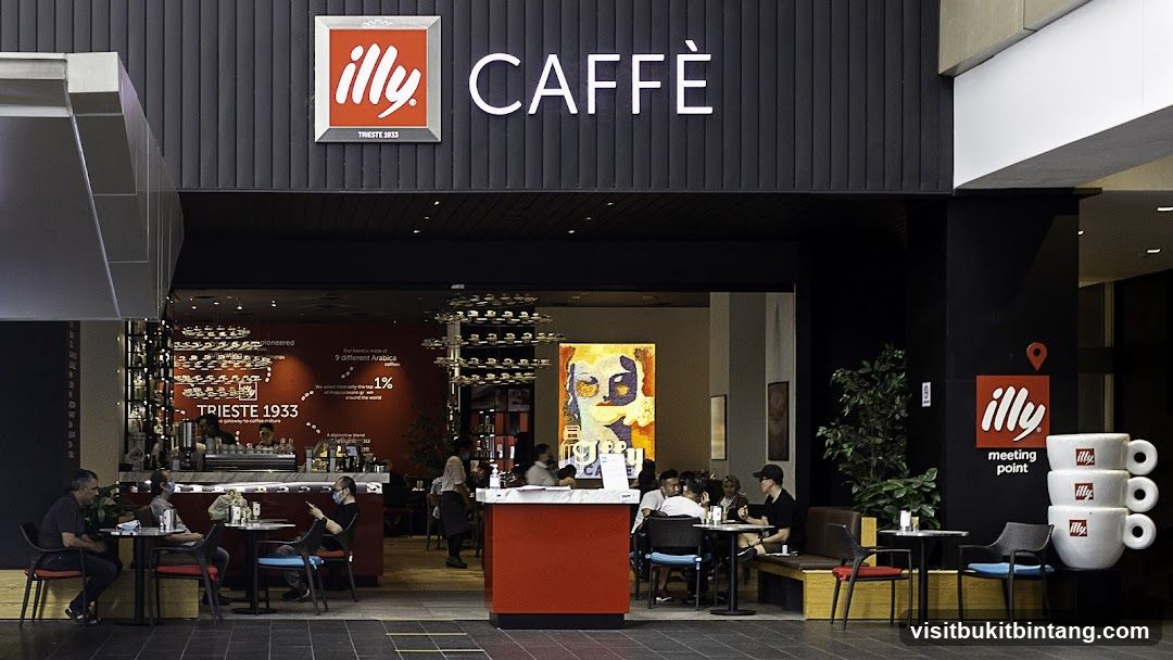 The exterior of an Illy Caffè shop with prominent signage. The shop features a modern design with black and red accents. Inside, customers are seated at tables, and a barista is visible behind the counter. A large artwork is displayed on the wall.