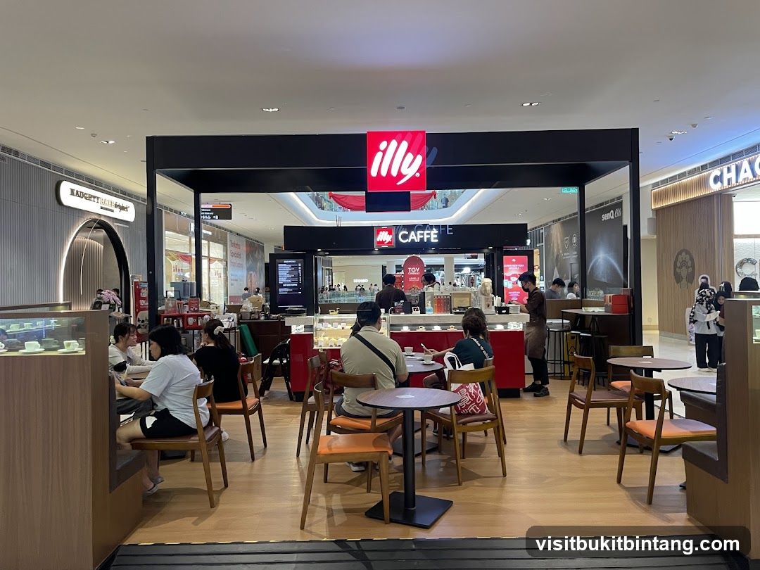 A view of the Illy Cafe storefront in a mall. The cafe has a black facade with the Illy logo in red and white. There are tables and chairs set up for customers inside and outside the cafe. People are seated at tables, and staff are visible behind the counter.