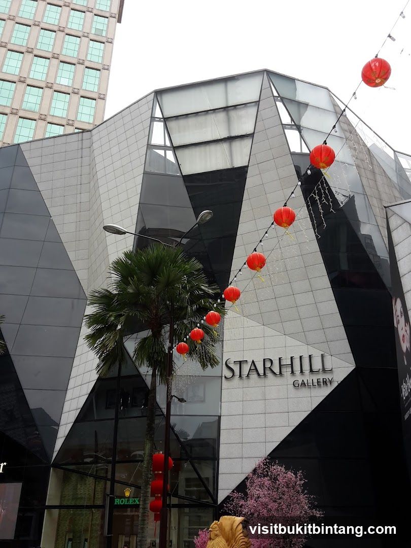 The exterior of a modern building with a geometric facade, featuring the signage "STARHILL GALLERY". Red lanterns are strung across the front, and a Rolex store is visible at street level. A palm tree stands in front of the building, and a decorative golden rooster and pink cherry blossoms are also present.