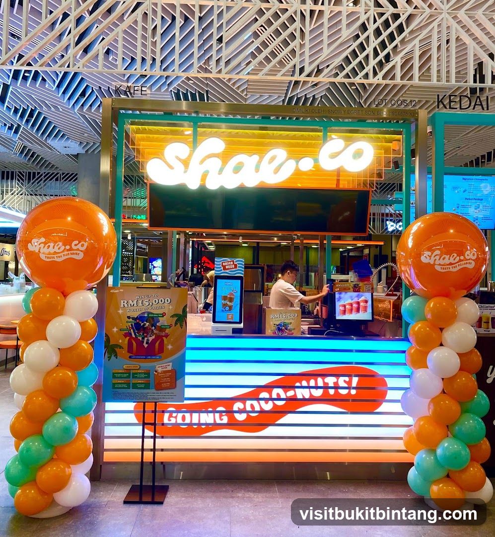 A brightly lit storefront for a beverage shop named Shae.co. The shop features a prominent neon sign with the business name. Two large orange balloons with the Shae.co logo are placed on either side of the entrance, and a colorful balloon arch decorates the left side. A promotional poster for a RM15,000 prize giveaway is displayed near the entrance. The counter area is visible with a staff member serving customers.