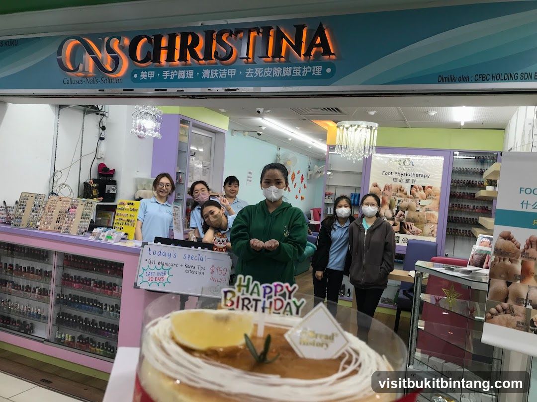 A group of women stand behind a counter in a nail salon. A cake with a "Happy Birthday" topper is in the foreground. The shop's name, "CNS CHRISTINA," is visible above the entrance. Displays of nail polish and nail art supplies are visible on shelves and in cabinets. A sign advertises "Foot Physiotherapy."