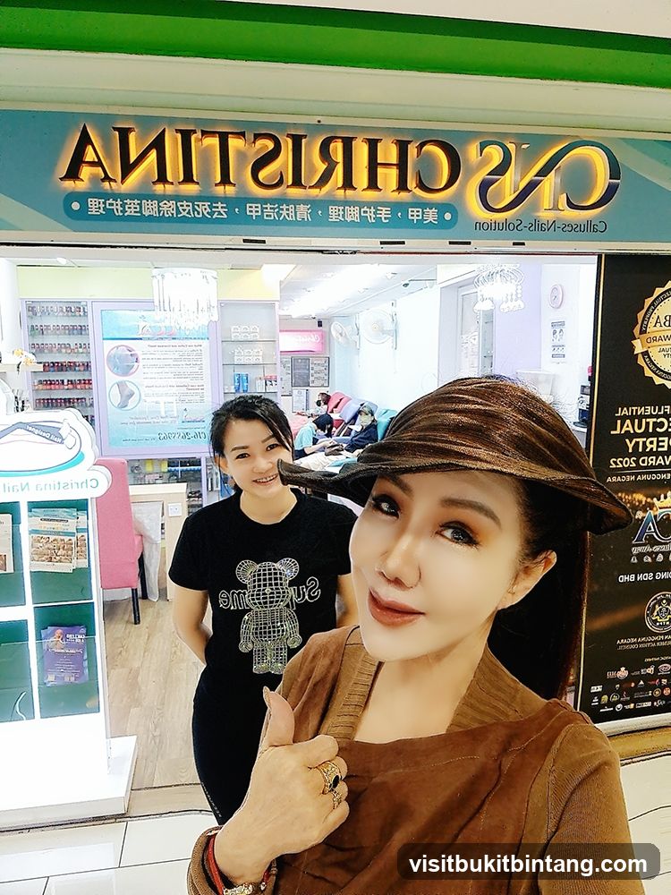 A woman in a brown cardigan and hat gives a thumbs up in front of a nail salon. Another woman stands behind her, smiling. The salon's sign, "CHRISTINA NAIL SOLUTION," is visible above them. Inside the salon, nail technicians can be seen working on clients.