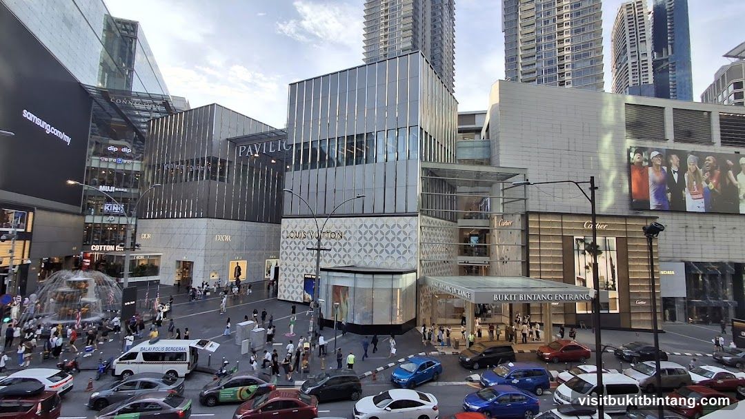 A wide shot of a busy street in front of the Pavilion Kuala Lumpur shopping mall. The mall features multiple luxury brand storefronts, including Louis Vuitton, Dior, and Cartier. The 'Pavilion' sign is visible on the building. The street is filled with cars and pedestrians. A large Samsung advertisement is displayed on a digital billboard.