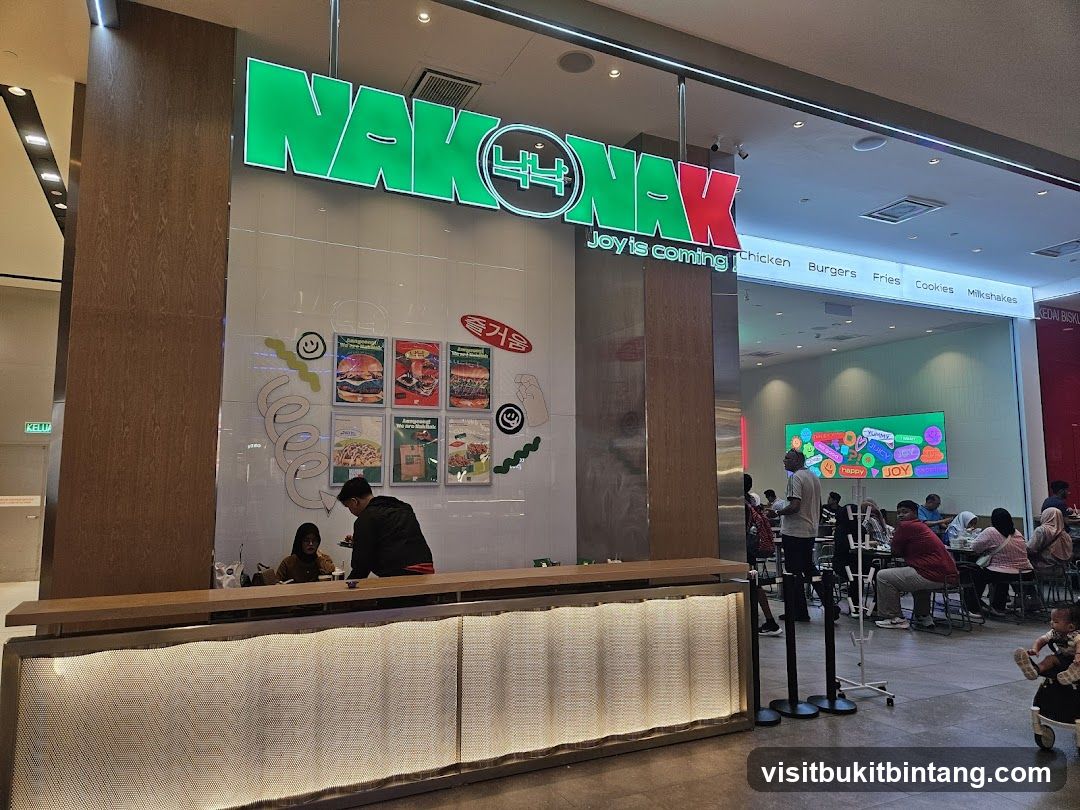 The exterior of a restaurant with a large green and red sign that says "NAK NAK" and "Joy is coming!". Below the sign, a menu board lists "Chicken, Burgers, Fries, Cookies, Milkshakes". Several framed posters with food images and Korean text are displayed on the wall. A counter with staff and customers is visible.