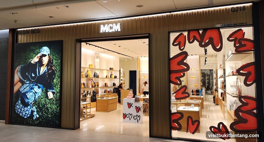 The exterior of an MCM store. The storefront features a large advertisement on the left, the MCM logo above the entrance, and a display window on the right decorated with red heart-like shapes. Inside, shelves are stocked with handbags and accessories.