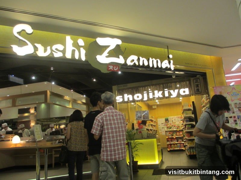 The exterior of a Sushi Zanmai Shojikiya restaurant. A large illuminated sign above the entrance reads "Sushi Zanmai". Below this, another sign reads "shojikiya". People are standing in line to enter. Inside, staff in white uniforms are visible behind a counter. Shelves stocked with products are visible to the right of the entrance.
