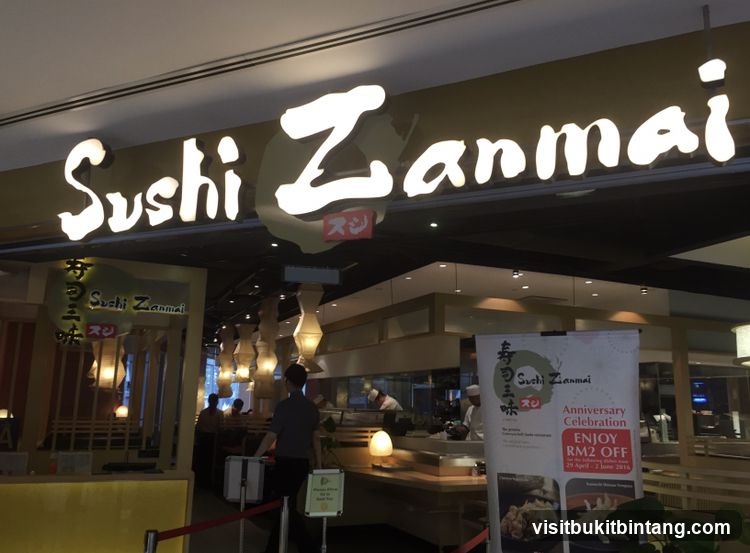 The exterior of a Sushi Zanmai restaurant. A large illuminated sign with the restaurant's name is visible above the entrance. Inside, diners can be seen at tables, and staff are working behind a counter. A promotional banner for an anniversary celebration offering RM2 off is displayed near the entrance.
