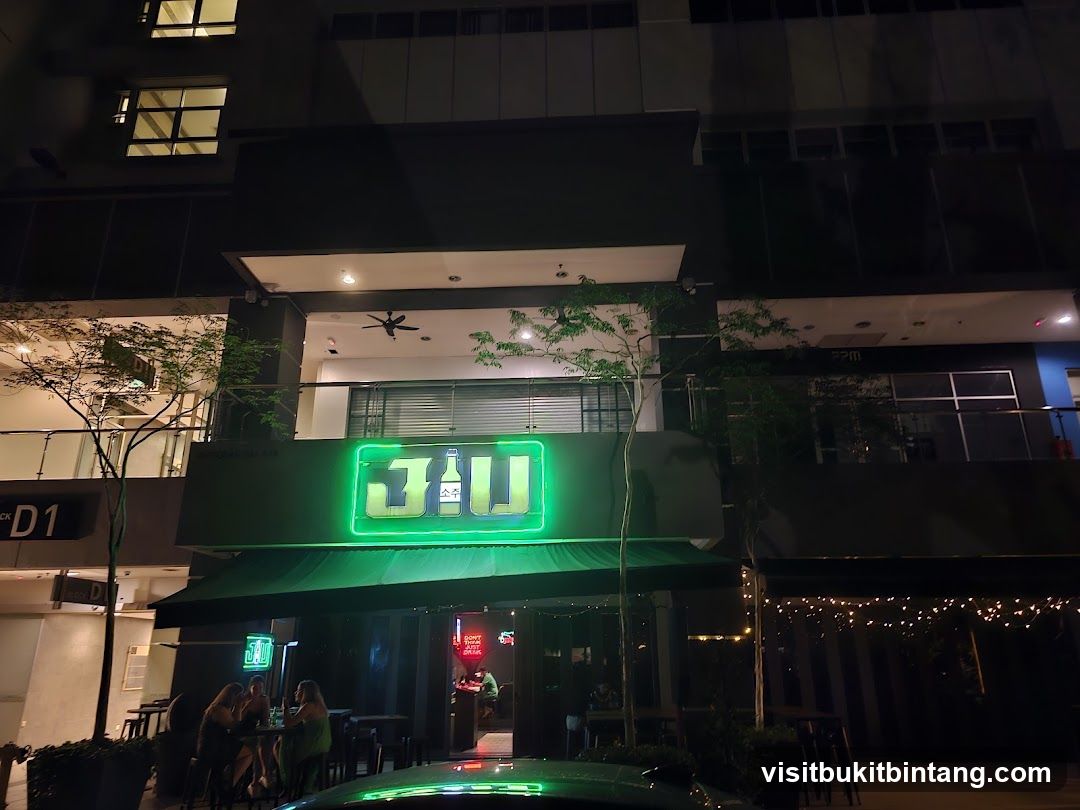 A neon green sign with the text "3JU" and Korean characters is illuminated above the entrance of a bar. The bar has a green awning and outdoor seating with people visible. The building has multiple floors with windows.