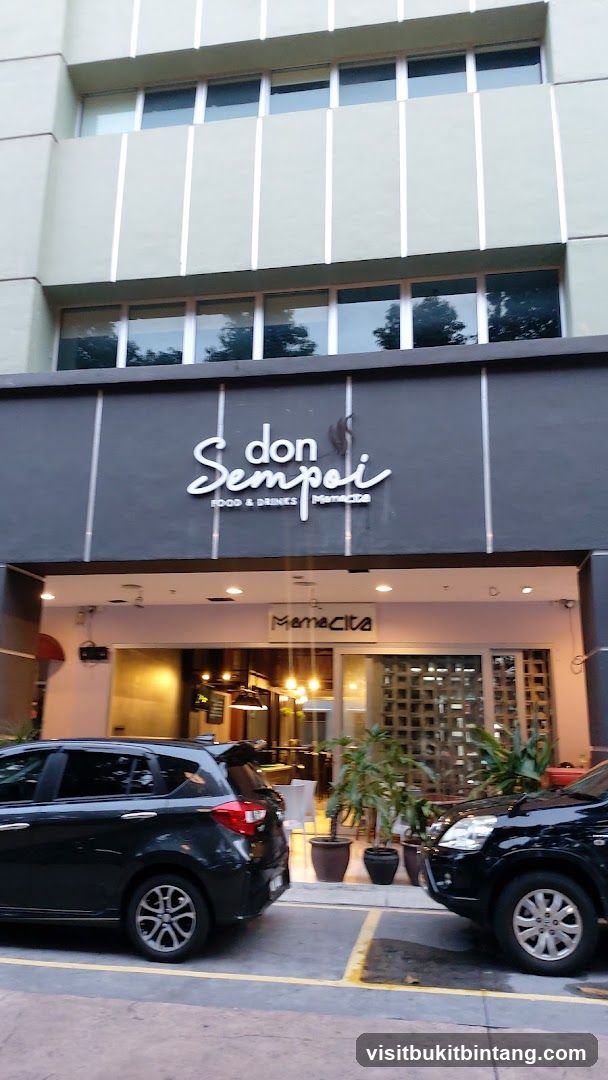 The exterior of a restaurant called Don Sempoi Food & Drinks. The name is displayed in a white, cursive font on a dark gray facade. Below this, the word "Mamacita" is visible on a smaller sign. The entrance is visible with glass doors and windows, and interior lighting can be seen. Two cars are parked in front of the establishment.