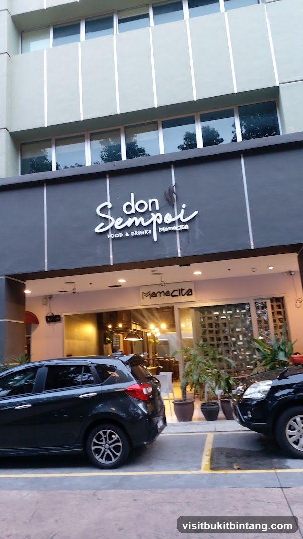 The exterior of a restaurant with a dark gray facade. The restaurant name "don Sempoi" is displayed in white cursive lettering above "FOOD & DRINKS" in smaller white text. Below this, the word "Mamacita" is also visible. The entrance to the restaurant is open, revealing a warmly lit interior with tables and chairs. Two cars are parked in front of the establishment.