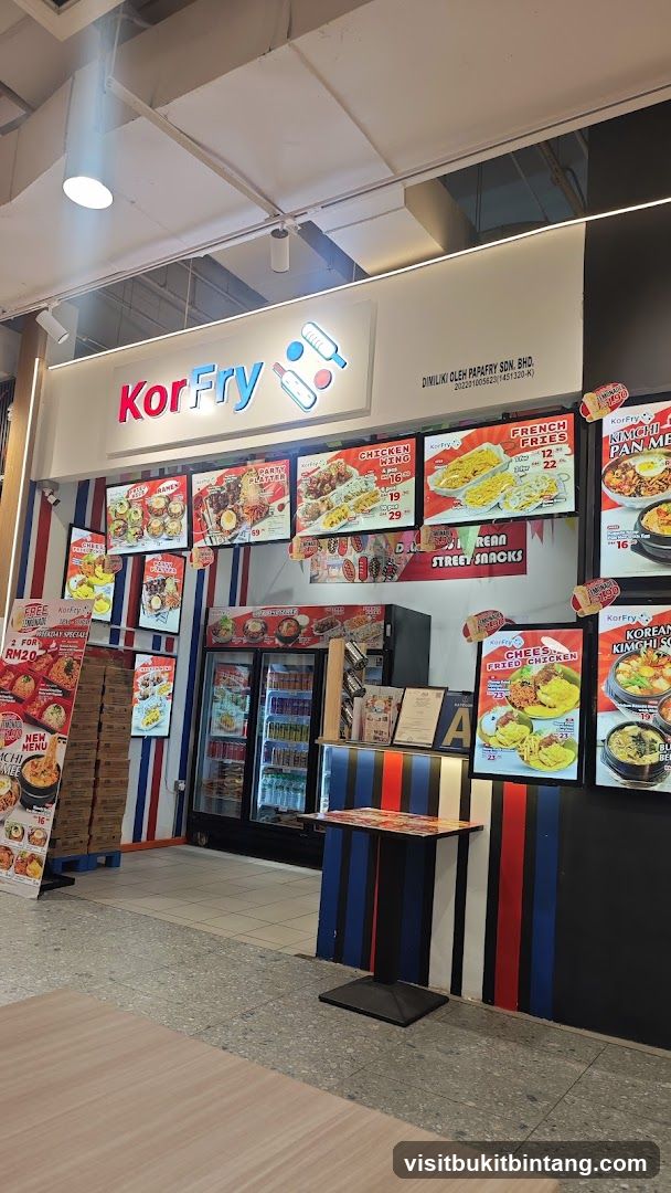 The exterior of a KorFry restaurant with its prominent signage and various food displays. The shop front features a large "KorFry" logo above the entrance. Digital menu boards showcase images and prices of Korean street food items like fried chicken, french fries, and kimchi ramen. A refrigerator stocked with drinks is visible behind the counter. The walls are decorated with vertical stripes in red, white, and blue.