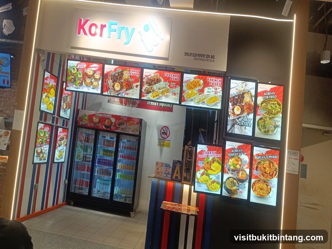 A brightly lit food stall with a prominent "KorFry" sign. The stall features multiple illuminated menu boards displaying various Korean-inspired dishes like fried rice, ramen, chicken wings, party platters, French fries, Korean stir-fried dishes, cheesy fried chicken, Korean kimchi soup, and Korean topokki. A large refrigerator stocked with drinks is visible behind the counter.