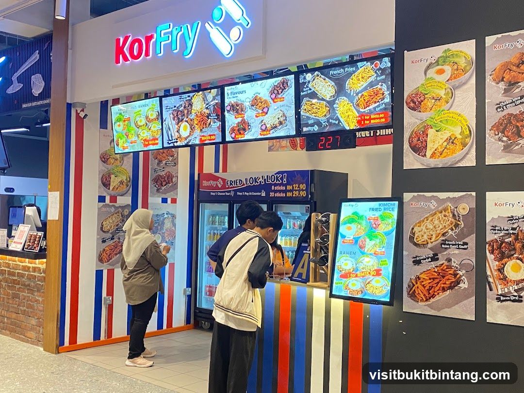 A KorFry food stall with a bright neon sign and digital menu boards displaying various Korean fried chicken and fries options. People are ordering at the counter.