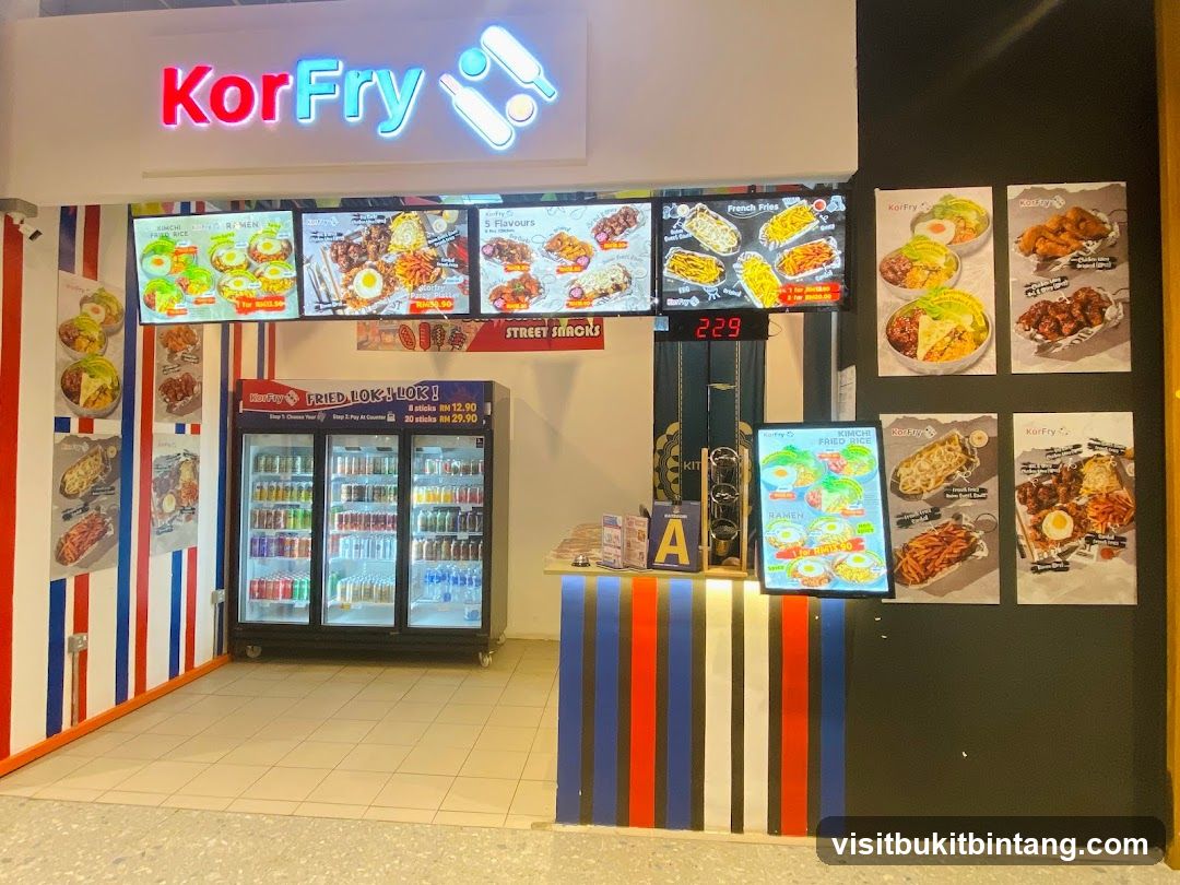 A KorFry store with a prominent sign, digital menu boards displaying food items and prices, a refrigerated display case filled with drinks, and promotional posters on the wall. The store features a colorful striped design on its facade.