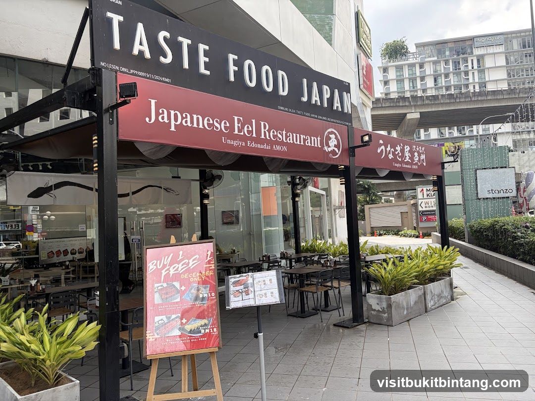 The exterior of a Japanese eel restaurant with outdoor seating. The restaurant has a large black sign with white lettering that reads "TASTE FOOD JAPAN" and a red banner below that says "Japanese Eel Restaurant" and "Unagiya Edonadai AMON". There are tables and chairs set up outside, and potted plants in front of the building. A promotional poster is displayed on an easel.