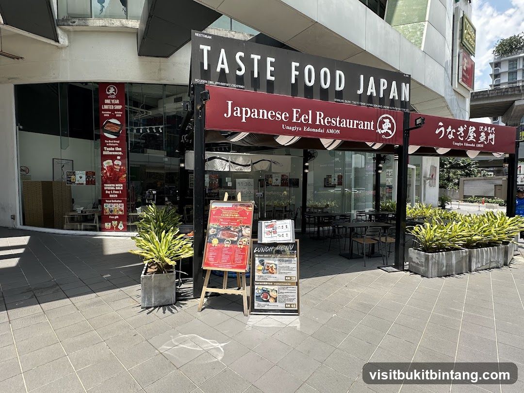 The exterior of a Japanese eel restaurant called Taste Food Japan, also known as Unagiya Edonadai AMON. The storefront features large glass windows and a prominent black and red awning with the restaurant's name and Japanese characters. A red banner on the left advertises a "One Year Limited Shop" and a "Tomato Beer Special Sale". Several potted plants are placed outside the entrance. A menu board is displayed on an easel in front of the restaurant.