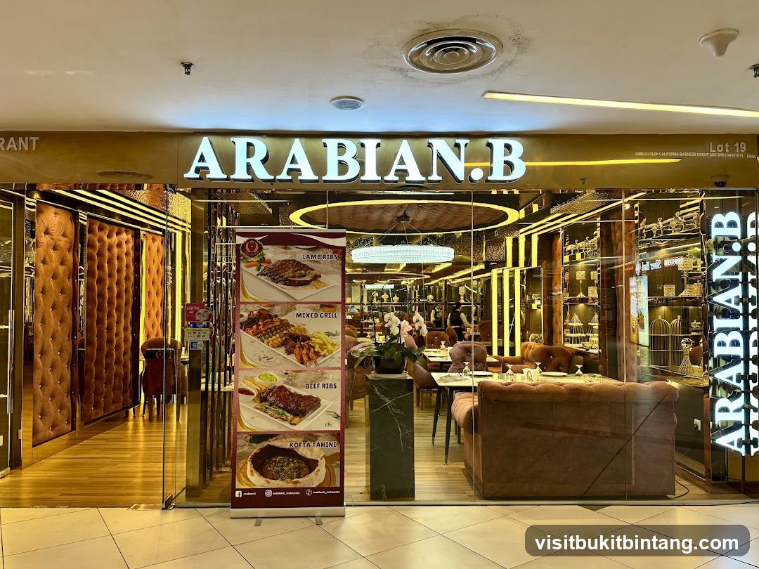 The exterior of a restaurant with a large illuminated sign that reads "ARABIAN.B". The entrance features glass doors and windows, revealing a well-lit interior with plush seating and decorative elements. A vertical banner displays images of various dishes, including Lamb Ribs, Mixed Grill, Beef Ribs, and Kofta Tahini.