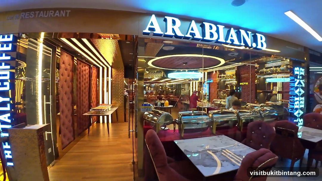 The exterior of a restaurant with a large sign that reads "ARABIAN.B" in illuminated white letters. To the left, a vertical neon sign reads "ITALY COFFEE". The entrance features glass doors and is flanked by plush, tufted red walls. Inside, a buffet setup with chafing dishes is visible, along with dining tables and chairs.