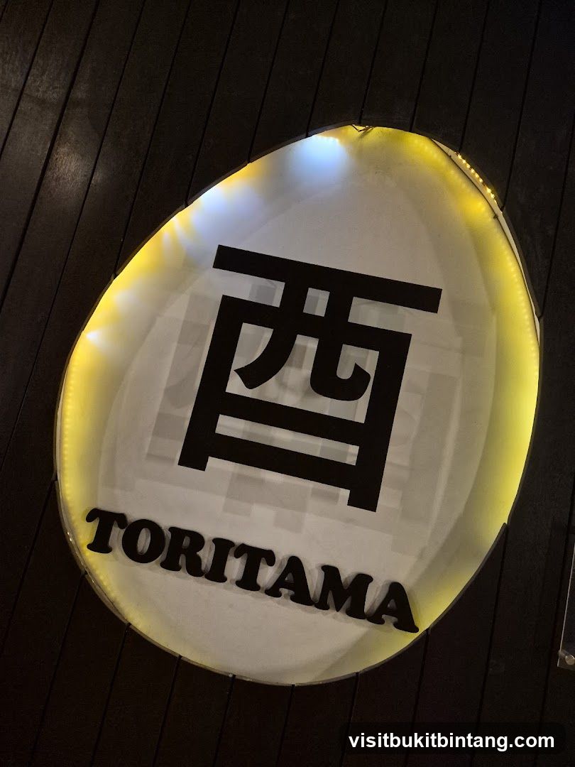 A close-up shot of a white, oval-shaped sign with a Japanese character and the word "TORITAMA" in black lettering. The sign is illuminated by yellow and white lights around its perimeter and is mounted on a dark wooden wall.